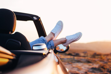 Trendy hipster woman rests and admire sunset in mountains from her cabriolet car. Happy female relaxing pushes her shoes out of convertible enjoy view