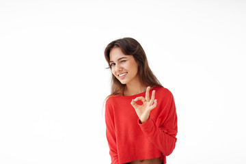 Wall Mural - Everything is just fine. Attractive joyful young Latin woman wearing red casual top making ok sing and winking at camera, having good mood, enjoying success or happy day, posing at studio wall