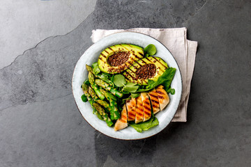 Healthy grilled chicken, grilled avocado and asparagus salad with linen seeds. Balanced lunch in bowl. Gray slate background. Top view