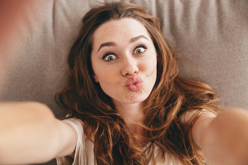 Wall Mural - Funny pretty lady lies on sofa indoors make selfie.