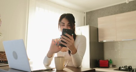 Poster - Young Woman Using Cell Smart Phone Sitting At Table With Laptop At Home Happy Smiling Beautiful Girl Messaging Online In Morning Indoors Slow Motion 60