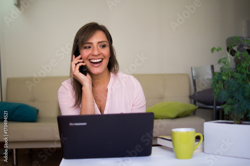 Mujer joven y bella hablando por teléfono frente al computador en la comodidad d Poster