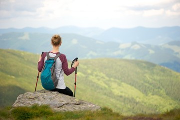 Short break on a hiking trip to enjoy the breathtaking view of the mountains. Young woman sitting on a rock, facing away from the camera; small backpack on her back and holding trekking sticks. 