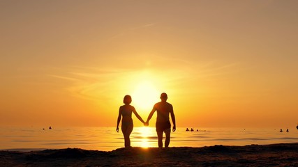 Happy couple walking along a sea beach holding hands on a sunset background