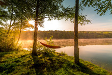 Summer Camping On The Lake. Empty Hammock  Between Two Trees With The View Of A Foggy Mountain Lake In Sunrise Light. Outdoors And Adventure Concept.
