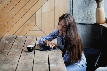 Beautiful cute Caucasian young female with beautiful long black hair in rustic cafe, using digital tablet and drinking coffee