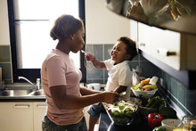 Black Kid Feeding Mother With Cooking Food In The Kitchen