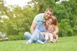 © serhiibobyk - Happy young man smiling joyfully embracing his beautiful wife and daughter sitting on the grass together copyspace family love emotions weekend enjoyment affection parents marriage.