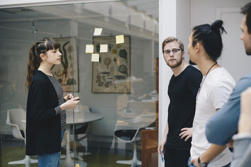 Male and female programmers discussing over adhesive notes stuck on glass in office