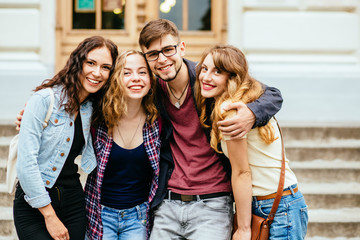 Cute group of four teenages at the building of university huggings, smiling, back to school concept.