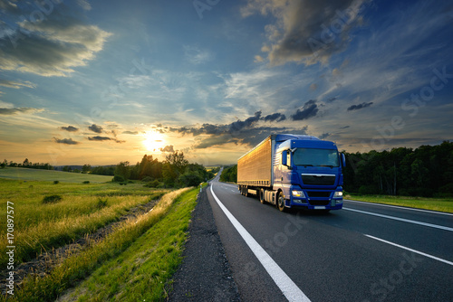 blue-truck-driving-on-the-asphalt-road-in-rural-landscape-at-sunset