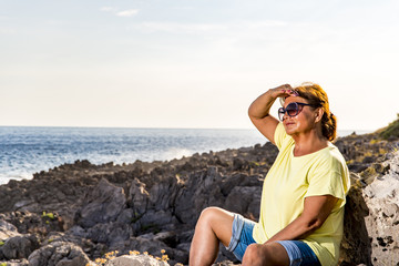 Dreamy beautiful woman in sunglasses with dark hair, dressed in shorts, sitting on large stones. Rocks on the background.