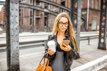 Lifestyle portrait of a stylish business woman with smartphone and coffeecup walking outdoors on the old iron bridge