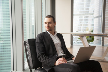 Confident handsome dreamy businessman holding laptop on his lap and looking towards window. Young CEO daydreaming, entrepreneur envisioning successful corporate future, profitable financial project