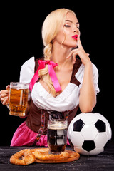 Wall Mural - Young sexy Oktoberfest girl - waitress, wearing a traditional Bavarian dress, serving big beer mugs and taking soccer ball on black background.