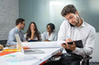 © Bojan - Young businessman holding mobile phone between ear and shoulder talking and writing notes to clipboard in modern office, while his business partners having meeting in the background.