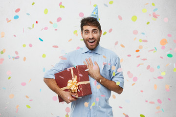 Horizontal shot of excited happy adult male holding gift box and keeping hand on his chest, touched with sincere cordial toast made by friend on his birthday party. Joy, happiness amd celebration