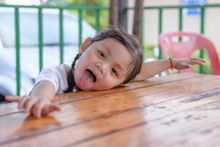 Happy Cute Asian Baby Girl In White T-shirt Stick Tongue Out And Put Her Face On Wooden Table.