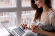 © Photodrive - Smiling young woman browsing laptop with a cup of hot drink at the window. Social life and remote working concept
