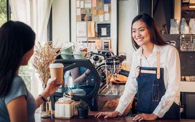 asian woman barista wear jean apron served to go coffee cup to customer at bar counter with smile em