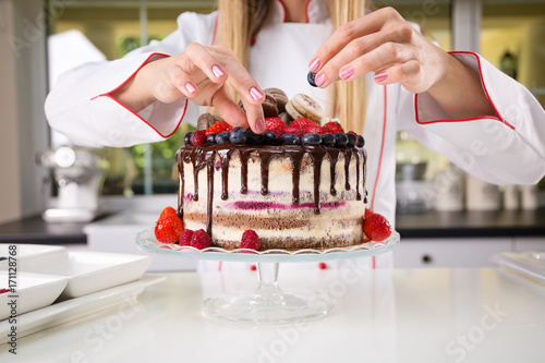 Plakat Młody żeński fachowy piekarz dekoruje czekoladowego i kremowego nagiego tort z czarnymi jagodami