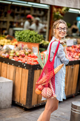 Wall Mural - Young woman standing outdoors with mesh bag full of fresh vegetables in front of the food market