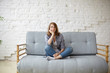 Portrait of beautiful pensive young Caucasian female keeping legs crossed while sitting on couch against white brick wall background, having thoughtful or bored look, dreaming of something pleasant