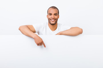 Cheerful young african man is standing behind the white blank banner and pointing down at a white copyspace, in casual wear