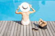 Beautiful young woman in hat relaxing near the swimming pool sitting back with bag and slippers on the wooden poolside