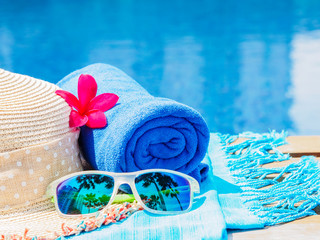 Wall Mural - Red frangipani (plumeria) flowers, sunglasses, beach hat and blue towel at the side of swimming pool. Vacation, beach, summer travel concept