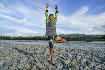 Young boy doing tree pose yoga on the shore of the beautiful mountain river.