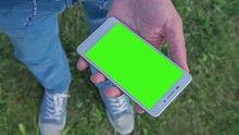 Young Man Staying In City Park Holds SmartPhone With Pre-keyed Green Screen. Perfect For Screen Compositing. Made From 14bit RAW. 10bit ProRes 444