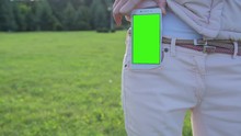 Young Woman Staying In City Park Pulls White SmartPhone With Pre-keyed Green Screen Out Of Her Pocket. Perfect For Screen Compositing. Made From 14bit RAW. 10bit ProRes 444