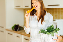 Portrait Of Charming Woman In Pajamas Standing In The Kitchen, Biting Carrot. Healthy Eating Concept. Diet.