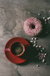 Pink glazed donut with pink sugar and red cup of black coffee over gray texture background. Flat lay with space