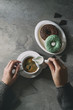 Plate of colorful glazed donuts with chopped chocolate, cup of black coffee, jug of milk over gray texture table. Female hands pouring cream. Flat lay with space. Rustic style