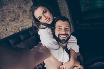 Sexy young cheerful brunette lady in white shirt looks at the camera, her handsome brunet bearded lover with beaming smile is piggy backing his lover, making selfie on his phone`s camera