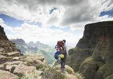 A Male Hiker With His Backpack Standing At A Mountain Cliff Edge Over A Mountainous Green Valley With Clouds.