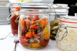 Glass preserving jars of pickled chillies, onions and gherkins on white table.