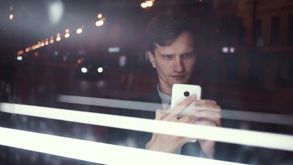Handsome young business man using smart phone on a busy urban street at night. Portrait of wearing casual man through light box. Car traffic at background. Slow motion