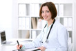 © rogerphoto - Young brunette female doctor sitting at the table and working at hospital office.