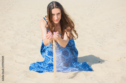 Photographie A woman with long dark hair, dressed in oriental style, in a blue dress with ornament sitting in the desert on the sand