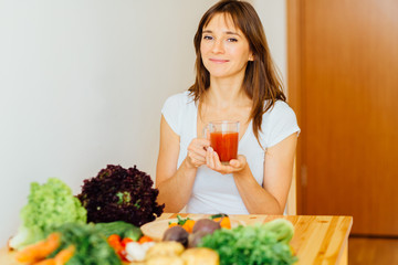 Healthy brunette woman drinking fresh detox Jjuice, Smoothie for breakfast. Closeup of beautiful smiling girl with vegetables at kitchen table. Nutrition concept.