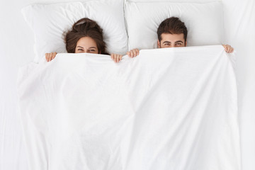 funny married couple lying in bed and hiding under white blanket, looking at camera with eyes full o