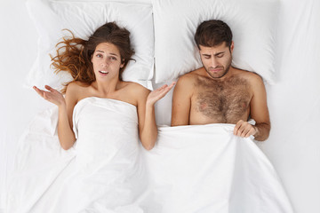 Frustrated young male impotent has problems with man`s health, looks under blanket at his organ, can`t satisfy wife in bed. Unhappy woman and male experience difficulties with personal relations