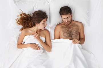 Relationships difficulties, stress and impotence. Couple having marital problems. Woman and man in bed looking under blanket, feeling unhappy and frustrated while husband having erectile dysfuntion