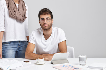 Wall Mural - Confident young bsinessman with earring and stubble studying documents at office with mug of coffee, electronic gadgets and papers on desk, his unrecognizable female assistant standing behind him