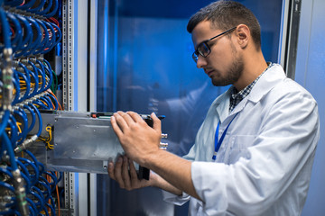 Side view portrait of young man in lab coat taking out blade server out of cabinet while working with supercomputer