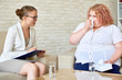 © Seventyfour - Profile view of confident young psychologist taking notes on clipboard while consulting crying obese woman during therapy session at cozy office
