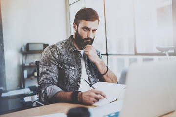 Wall Mural - Bearded young businessman working at sunny office.Man using contemporary laptop and making notes in notebook.Horizontal.Blurred background.